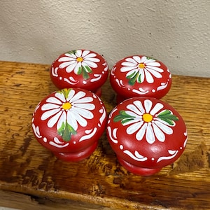May include: Four red cabinet knobs with white daisy designs and green leaves. The knobs are round with a slightly flared base, and are arranged on a wooden surface. The daisy petals are white with yellow centers.
