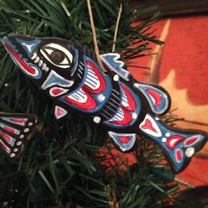 May include: A hand-painted wooden ornament shaped like a fish with red, blue, white, and black details. The ornament is hanging from a green artificial Christmas tree.
