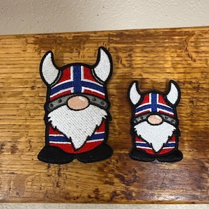 May include: Two embroidered patches featuring Viking gnomes. The gnomes have white beards, red and blue hats, and silver accents. The patches are on a wooden surface.