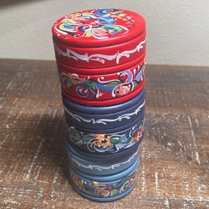 May include: A stack of four small, round, wooden boxes. The top box is red with a floral design on the lid. The other boxes are blue, with white and floral accents. The boxes are stacked on top of each other.