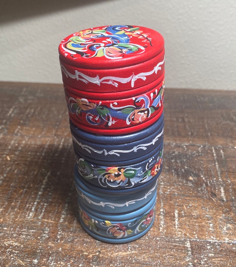 May include: A stack of four small, round, wooden boxes. The top box is red with a floral design on the lid. The other boxes are blue, with white and floral accents. The boxes are stacked on top of each other.