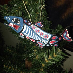 May include: A blue, red, and white painted wooden salmon ornament with a string for hanging. The salmon is stylized with geometric patterns and circles.