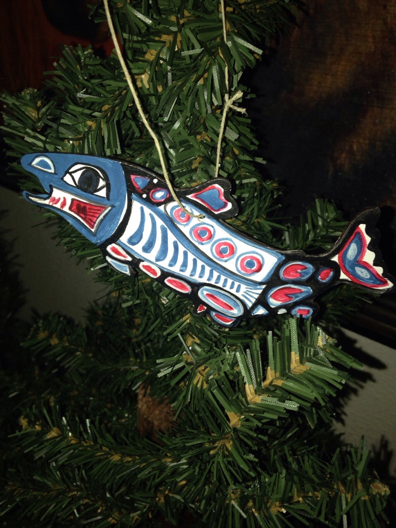 May include: A blue, red, and white painted wooden salmon ornament with a string for hanging. The salmon is stylized with geometric patterns and circles.