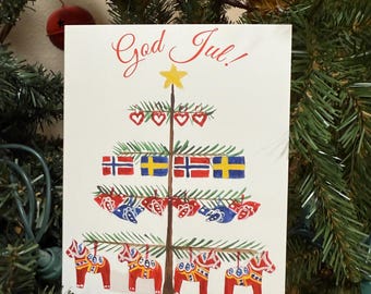 Op de afbeelding: Een kerstkaart met een boomontwerp, met vlaggen en Dala-paarden. De kaart zegt "God Jul!" in rood schrift. De boom heeft een gele ster bovenop en rode harten. De kaart wordt op een kerstboom weergegeven.