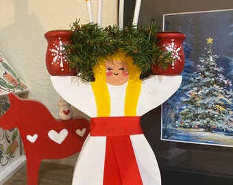 Puede incluir: Una figurita de ángel de madera blanca con un halo amarillo, una cinta roja y una corona de laurel verde en la cabeza. El ángel sostiene dos candelabros rojos con diseños de copos de nieve blancos.