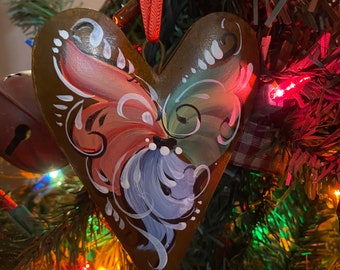 May include: A heart-shaped Christmas ornament with a brown metal base. The ornament features a hand-painted design with pink, blue, and white floral accents. A red ribbon is attached for hanging. The ornament is displayed on a Christmas tree.