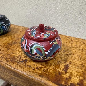 May include: A small, round, red wooden trinket box with a lid. The box and lid are adorned with colorful floral and abstract designs in blue, green, white, and yellow. The box sits on a wooden surface.