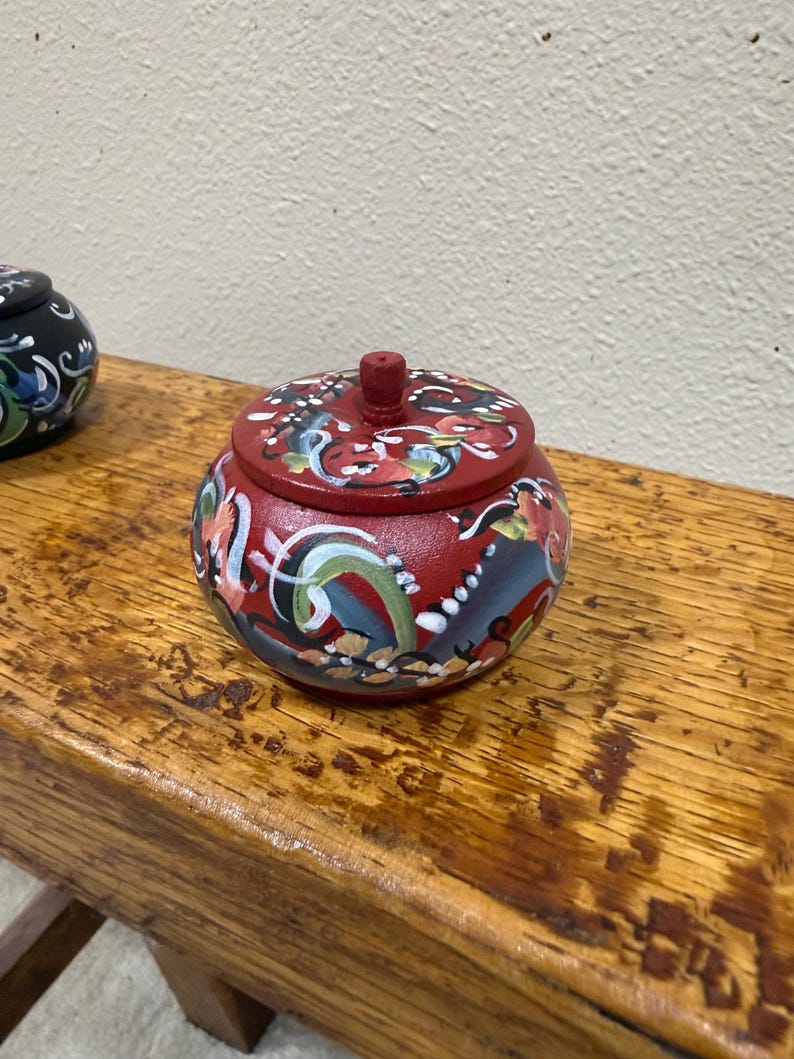 May include: A small, round, red wooden trinket box with a lid. The box and lid are adorned with colorful floral and abstract designs in blue, green, white, and yellow. The box sits on a wooden surface.