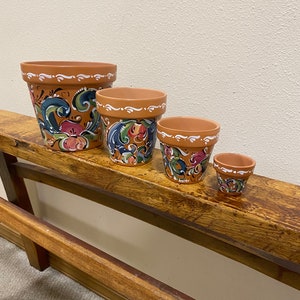 May include: Four terracotta flower pots with hand-painted floral designs in blue, green, pink, and white. The pots are arranged in a row from largest to smallest on a wooden shelf.