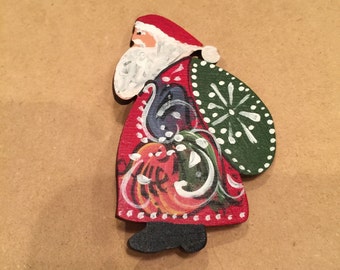 Op de afbeelding: Een handgeschilderd houten ornament van Sinterklaas met een zak vol cadeaus. Hij draagt een rood pak met witte versieringen, een witte baard en een rode muts. De zak is groen met witte sneeuwvlokken.