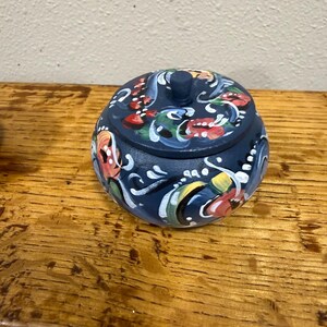 May include: A small, round, navy blue decorative box with a lid. The box is adorned with colorful floral and scroll designs in red, white, yellow, and green. The box is sitting on a wooden surface.