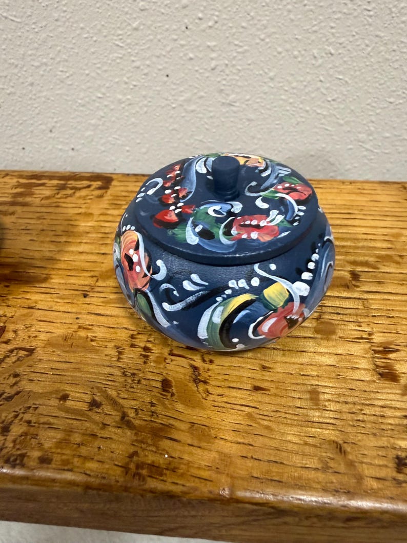 May include: A small, round, navy blue decorative box with a lid. The box is adorned with colorful floral and scroll designs in red, white, yellow, and green. The box is sitting on a wooden surface.