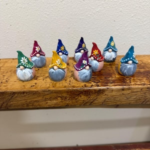 May include: A collection of nine small gnome figurines, each featuring a pointed hat in various colors including green, red, blue, yellow, and purple, decorated with painted white or yellow flower patterns and a long, textured gray beard.