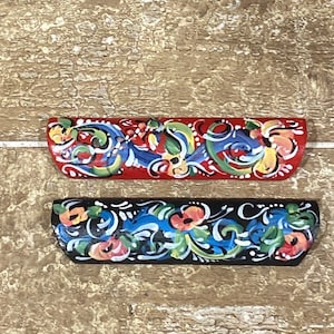 May include: Two painted wooden ornaments with a traditional folk art design. The top ornament is red with blue, green, and yellow floral patterns. The bottom ornament is black with blue, green, and orange floral patterns.