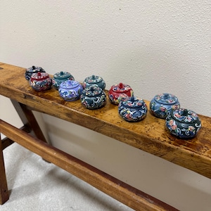 May include: A collection of small, round, lidded decorative boxes with floral designs. The boxes are painted in various colors, including blue, red, and green, and are arranged on a wooden surface. Each box has a small knob on top.