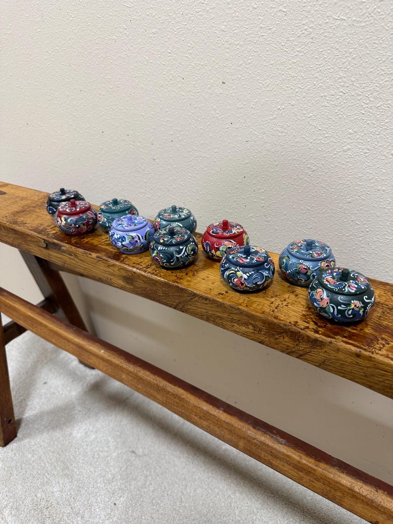 May include: A collection of small, round, lidded decorative boxes with floral designs. The boxes are painted in various colors, including blue, red, and green, and are arranged on a wooden surface. Each box has a small knob on top.