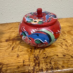 May include: A small, round, red wooden jar with a lid. The jar is decorated with colorful floral and abstract designs in blue, green, and white. The lid has a small, red knob. The jar sits on a wooden surface.