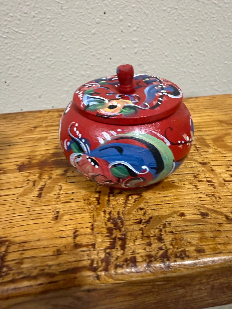 May include: A small, round, red wooden jar with a lid. The jar is decorated with colorful floral and abstract designs in blue, green, and white. The lid has a small, red knob. The jar sits on a wooden surface.