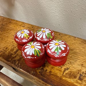 May include: Four red, wooden knobs with white daisy designs and yellow centers. The knobs are stacked on a wooden surface. The daisy petals are outlined in black, and the leaves are green. The knobs are a decorative accent.