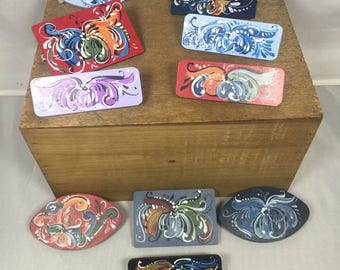 May include: A collection of eight hand-painted wooden ornaments featuring traditional Swedish folk art designs. The ornaments are painted in various colors, including blue, red, green, and yellow. They are all in the shape of a rectangle or oval.