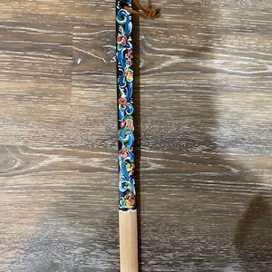 Op de afbeelding: Een houten stok met een zwart geverfd gedeelte met een bloemenmotief in blauw, rood, groen en geel. De stok heeft een bruine leren lus aan de bovenkant.