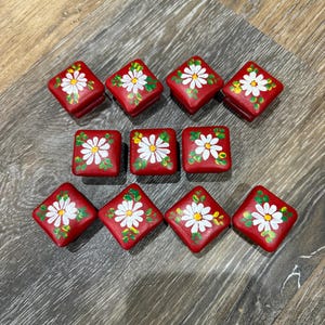 May include: Ten square wooden buttons painted in a vibrant red color. Each button features a hand-painted white daisy with a yellow center and green leaves. The buttons are arranged in three rows, with some positioned as squares and others as diamonds.