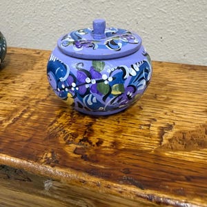 May include: A small, round, hand-painted wooden jar with a lid. The jar is painted in shades of blue and purple, with floral designs in white, yellow, and green. The jar sits on a wooden surface.