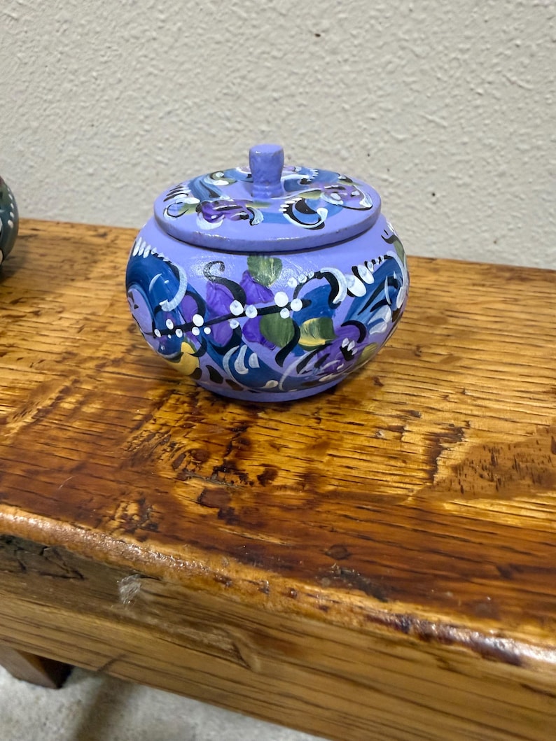 May include: A small, round, hand-painted wooden jar with a lid. The jar is painted in shades of blue and purple, with floral designs in white, yellow, and green. The jar sits on a wooden surface.