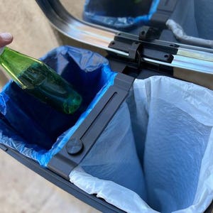 May include: A dual-compartment trash can with a stainless steel lid and a dark gray frame. One side has a blue trash bag, and the other has a white bag. A green bottle is being placed into the blue bag.
