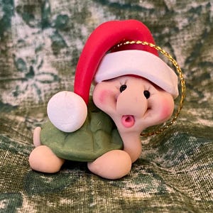 May include: A small, green turtle ornament wearing a red Santa hat with a white trim and a gold string for hanging. The turtle has a white snowball in its hand.