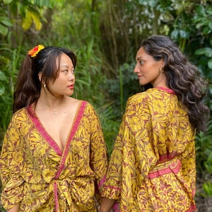 May include: Two matching wrap robes featuring a vibrant yellow and brown floral pattern with contrasting magenta trim along the V-neckline, sleeves, and waist tie. The robes have wide sleeves and a wrap-style front closure with a fabric belt.