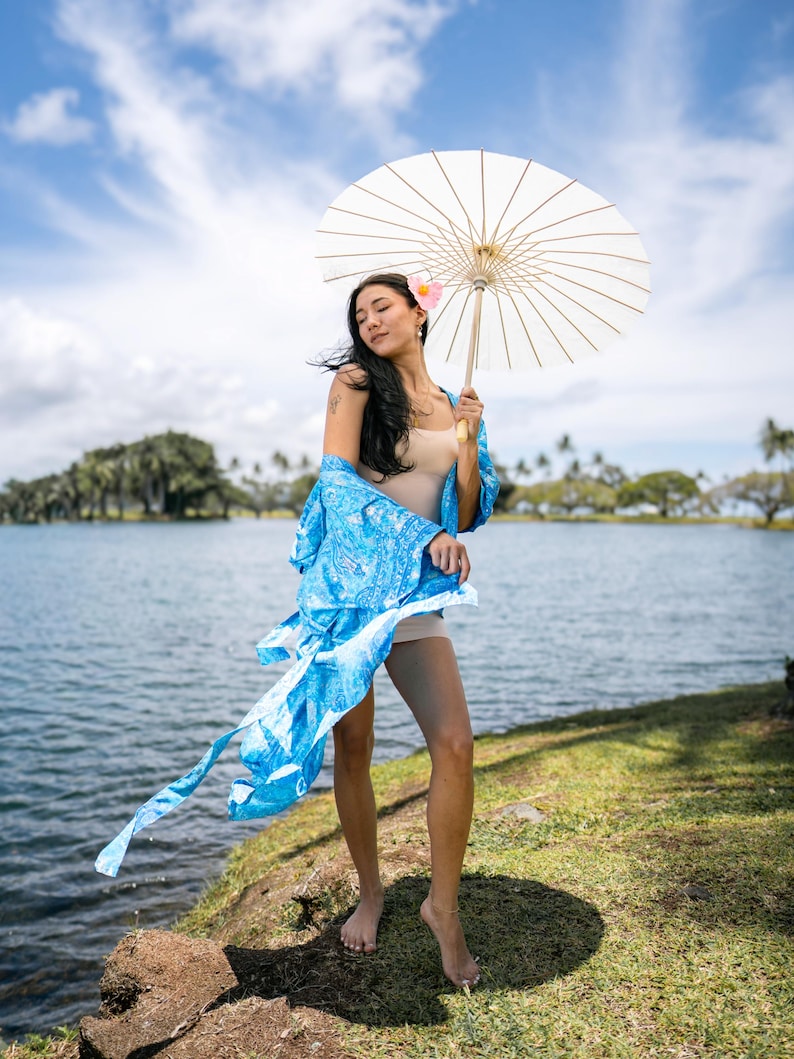 May include: A light blue robe with a white paisley pattern worn over a beige bodysuit, paired with a white paper parasol featuring a wooden handle and a pink flower tucked behind the ear, standing near a body of water under a blue sky.