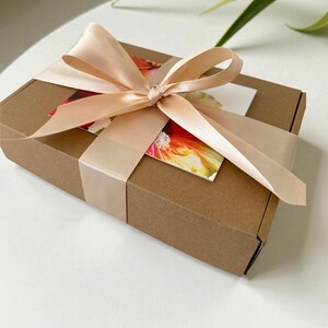 May include: A rectangular brown cardboard gift box wrapped with a wide, pale peach satin ribbon tied in a bow. A rectangular card featuring a vibrant, colorful floral print in shades of red, orange, and yellow is tucked beneath the ribbon.