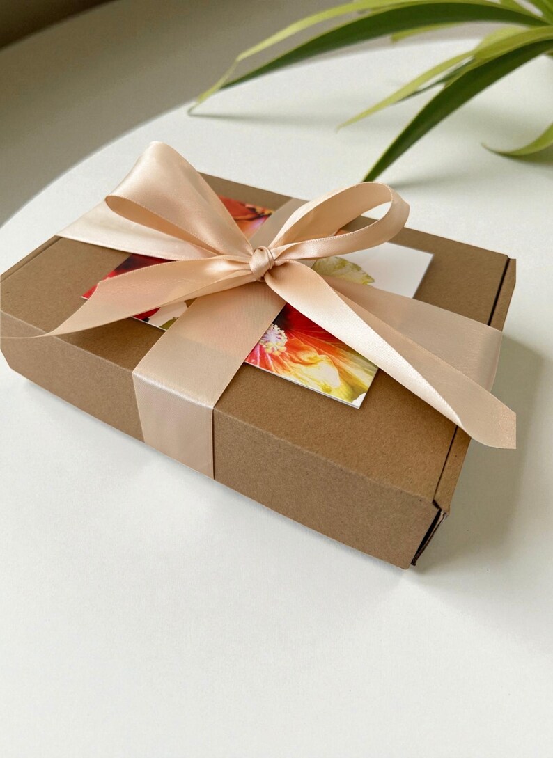 May include: A rectangular brown cardboard gift box wrapped with a wide, pale peach satin ribbon tied in a bow. A rectangular card featuring a vibrant, colorful floral print in shades of red, orange, and yellow is tucked beneath the ribbon.