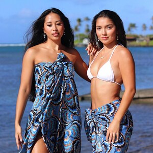 May include: Two women wearing beachwear by the ocean. One wears a blue and black patterned sarong as a dress. The other wears a white bikini top and a matching sarong. The sarongs have a blue, black, and orange design.