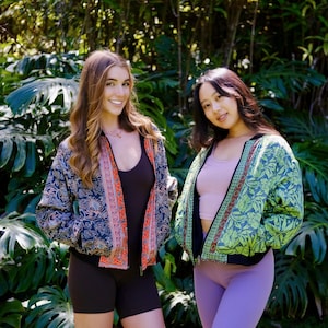 Può includere: Due donne indossano giacche bomber con motivi. La donna a sinistra indossa una canotta nera e pantaloncini neri. La donna a destra indossa una canotta rosa chiaro e leggings viola. Entrambe le donne sono in piedi davanti a uno sfondo verde lussureggiante.