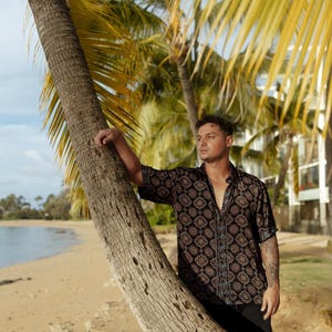 Puede incluir: Una camisa de manga corta con un patr&oacute;n geom&eacute;trico negro y marr&oacute;n. La camisa tiene un borde azul en la parte delantera y se usa con pantalones negros. El fondo incluye una playa, palmeras y un cielo azul.