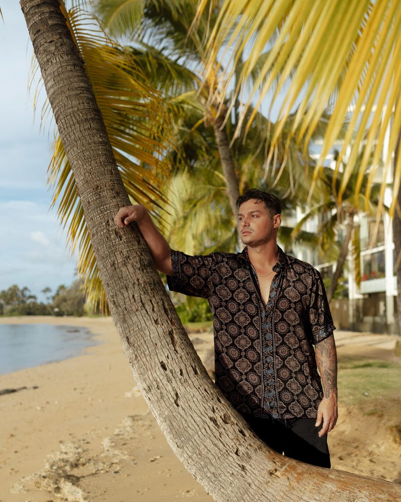 Puede incluir: Una camisa de manga corta con un patr&oacute;n geom&eacute;trico negro y marr&oacute;n. La camisa tiene un borde azul en la parte delantera y se usa con pantalones negros. El fondo incluye una playa, palmeras y un cielo azul.