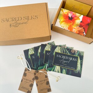 May include: A brown cardboard shipping box with the text Sacred Silks Apparel, an open box containing a floral card and a gold note, several rectangular cards with palm leaf prints and the text Sacred Silks Apparel, and two brown paper tags.