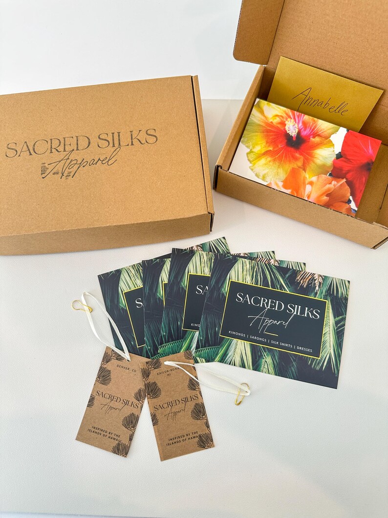 May include: A brown cardboard shipping box with the text Sacred Silks Apparel, an open box containing a floral card and a gold note, several rectangular cards with palm leaf prints and the text Sacred Silks Apparel, and two brown paper tags.