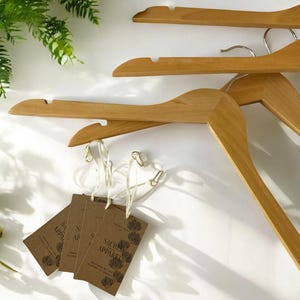 May include: Wooden clothes hangers with metal hooks and attached tags. The tags are brown with the words "Sacred Apparel" and a floral design. The hangers are light brown and are arranged on a white background.