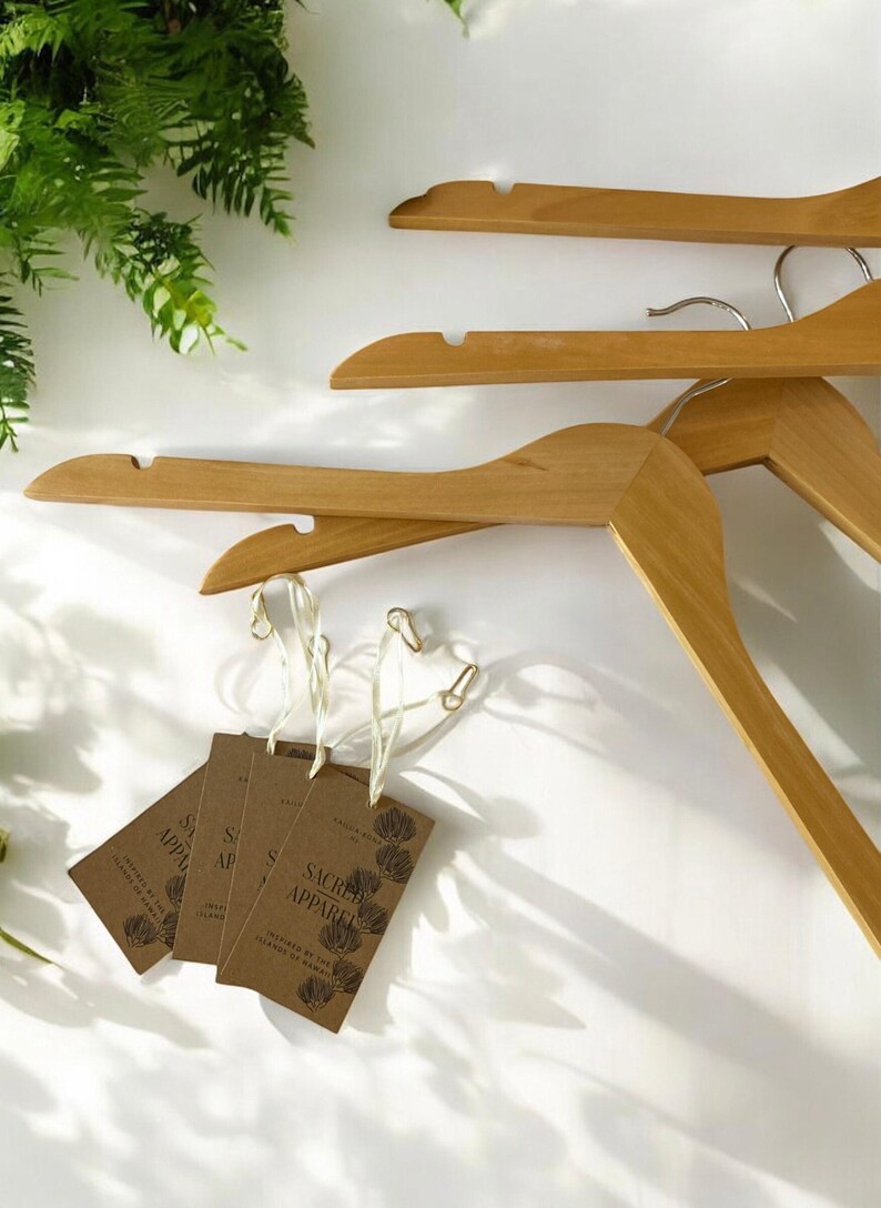 May include: Wooden clothes hangers with metal hooks and attached tags. The tags are brown with the words "Sacred Apparel" and a floral design. The hangers are light brown and are arranged on a white background.