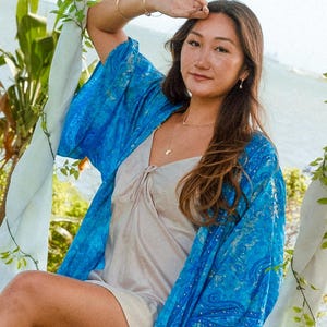 May include: A bright blue kimono featuring a detailed paisley pattern with subtle metallic gold accents, worn over a simple beige sleeveless slip dress with a V-neckline and a small tie detail at the center of the chest.