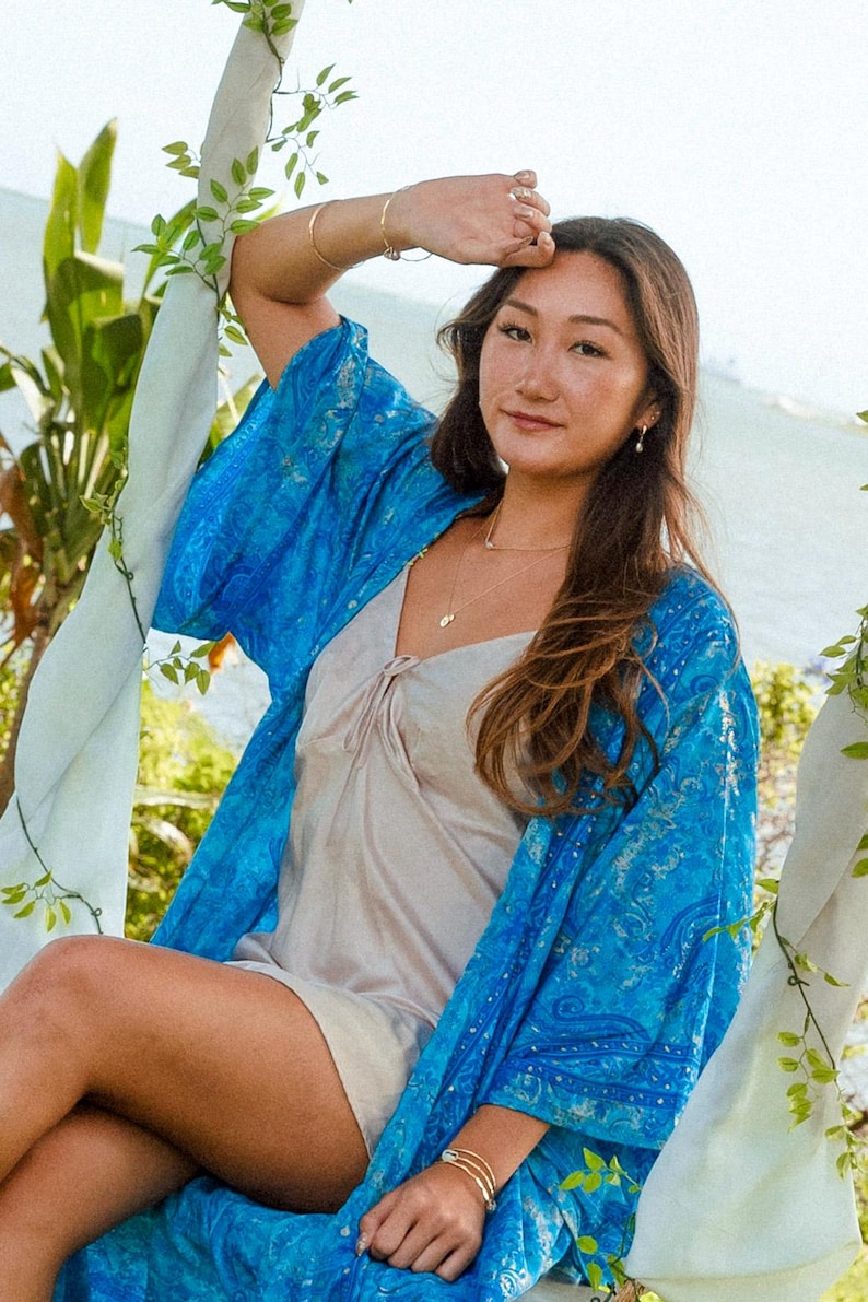 May include: A bright blue kimono featuring a detailed paisley pattern with subtle metallic gold accents, worn over a simple beige sleeveless slip dress with a V-neckline and a small tie detail at the center of the chest.