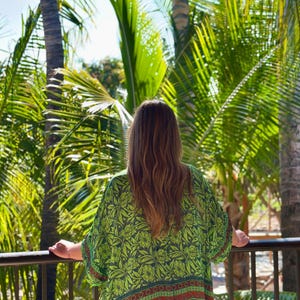 Puede incluir: Una persona con un kimono verde con estampado floral, de pie en un balc&oacute;n con vistas a palmeras. El kimono tiene una base verde oscuro con dise&ntilde;os florales verde claro y detalles rojos y naranjas. La persona est&aacute; de espaldas.