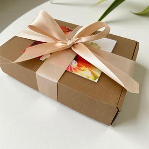 Pu&ograve; includere: Una scatola regalo rettangolare in cartone marrone avvolta da un ampio nastro di raso color pesca pallido annodato a fiocco. Sotto il nastro &egrave; inserito un cartoncino rettangolare con una vivace stampa floreale nei toni del rosso, arancione e giallo.