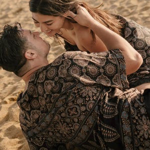 Puede incluir: Una pareja abraz&aacute;ndose en una playa arenosa. Llevan ropa negra estampada a juego con detalles en azul y beige. La mujer sonr&iacute;e y toca el pelo del hombre.