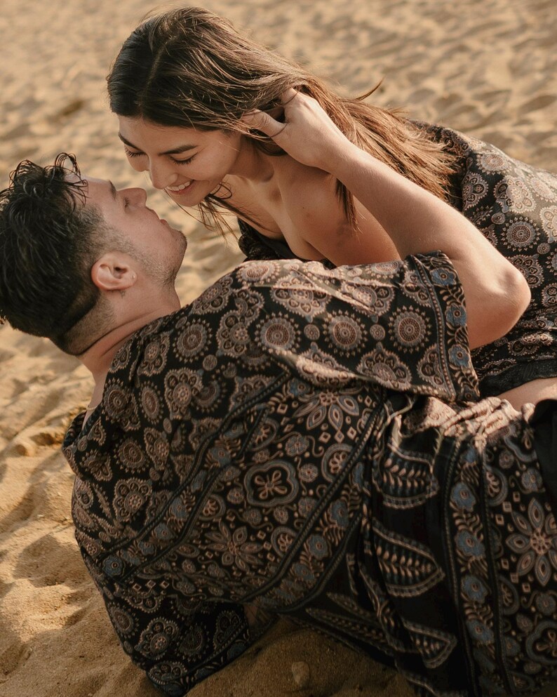 Puede incluir: Una pareja abraz&aacute;ndose en una playa arenosa. Llevan ropa negra estampada a juego con detalles en azul y beige. La mujer sonr&iacute;e y toca el pelo del hombre.