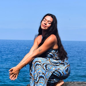May include: A blue and white patterned jumpsuit with a halter neckline. The jumpsuit features a vibrant design with orange and black accents. The person is squatting near the ocean, with the blue water and sky in the background.