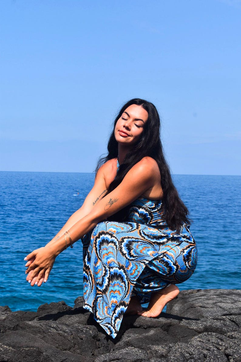 May include: A blue and white patterned jumpsuit with a halter neckline. The jumpsuit features a vibrant design with orange and black accents. The person is squatting near the ocean, with the blue water and sky in the background.